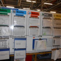A board showing daily notices for factory employees, including Health and Safety sheets and Risk Assessment binders 2013.